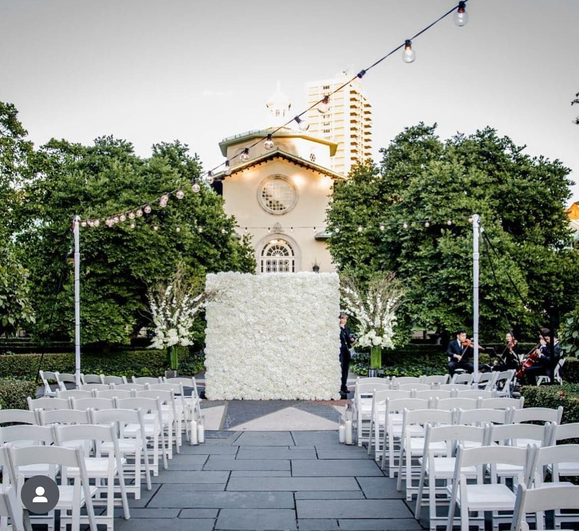 White flower wall backdrop at an outdoor wedding ceremony, string lights overhead